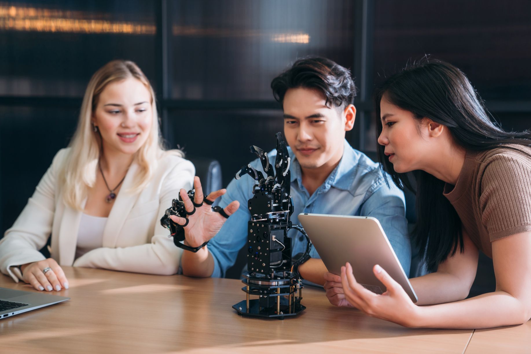 Three researchers testing a robotic hand prototype while reviewing the project on a tablet and laptop.